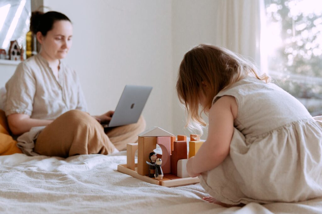 cute toddler girl playing with wooden toys on bed while mother using laptop